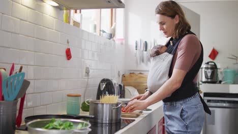 video of caucasian mother with newborn baby in baby carrier cooking in kitchen