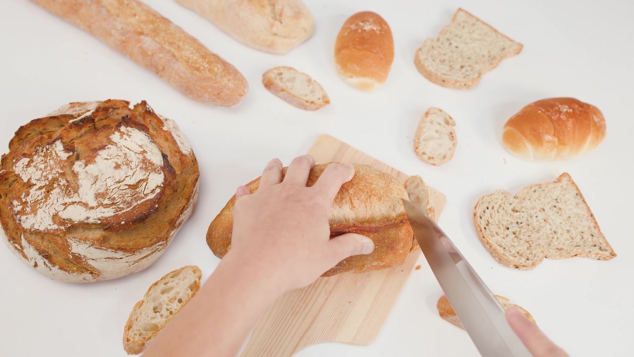 Ripresa dall'alto di un fornaio che affetta una pagnotta di pane a lievitazione naturale croccante su un tagliere di legno su sfondo bianco