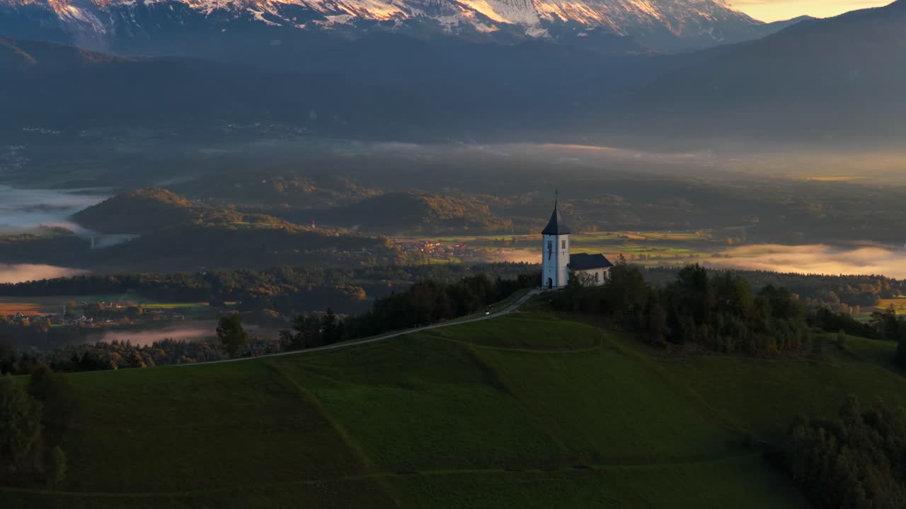 Aerial establishing shot of the Church of St. Primus and Felician on Jamnik Hill, Slovenia, overlooking a misty valley with the Julian Alps in the background during early morning light