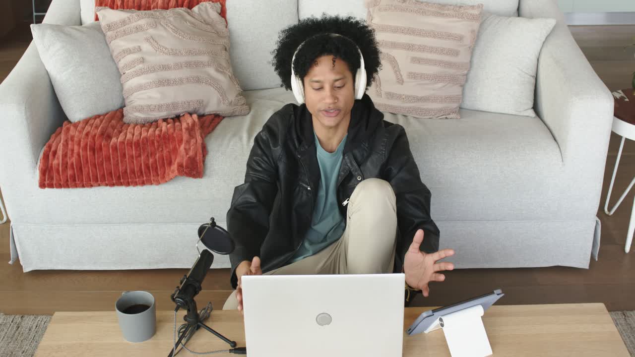 Afroamerikanischer Mann, der zu Hause einen Podcast aufnimmt, indem er den Laptop öffnet, die Kopfhörer einstellt und in ein Mikrofon spricht