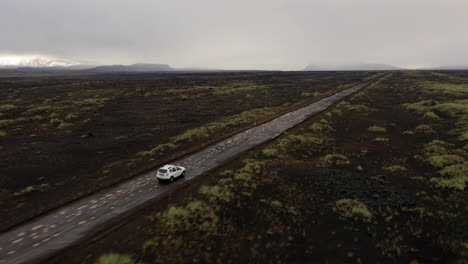 side aerial follow shot of white car driving on black gravel road amongst green moss fields in highlands of iceland