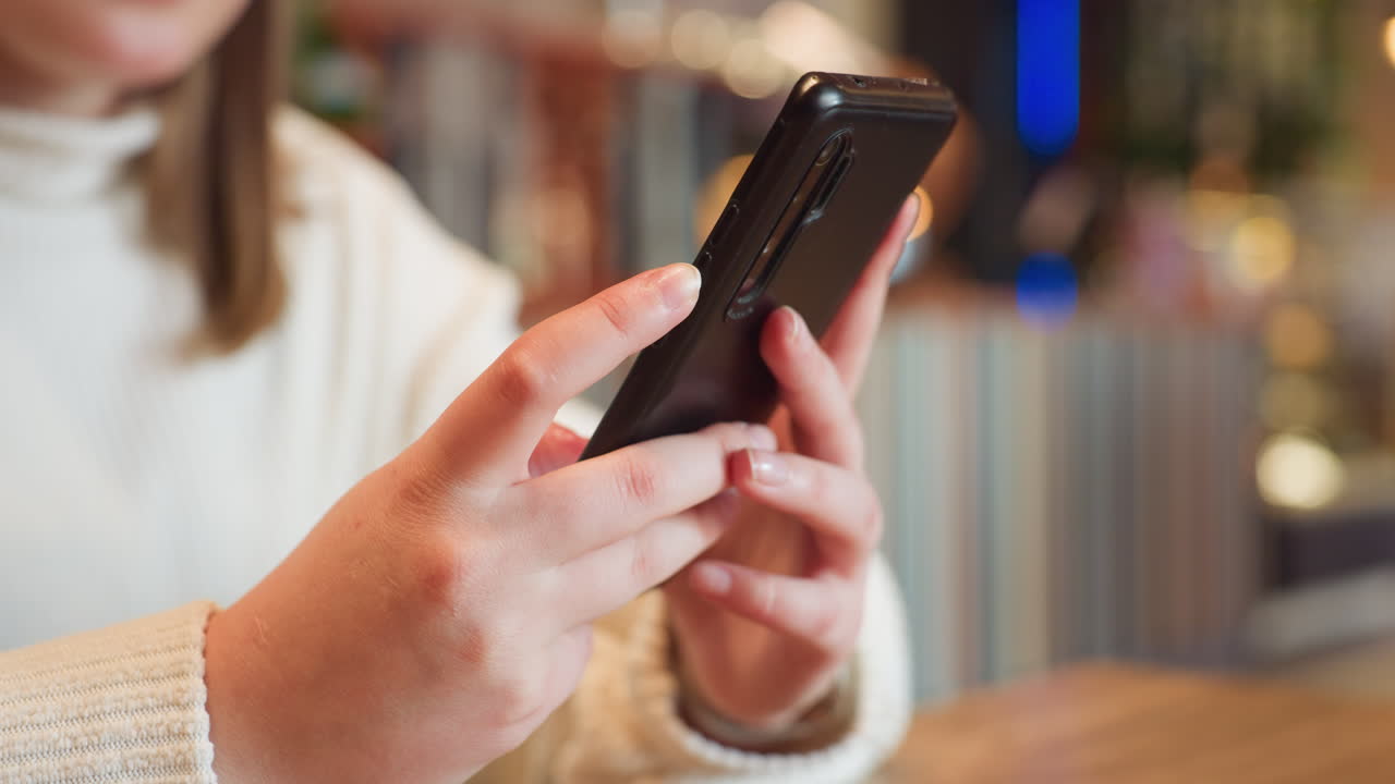 Close up of hands of young woman wearing fluffy white sweater operating smartphone indoors with fingers gently holding device, soft light and blurry background