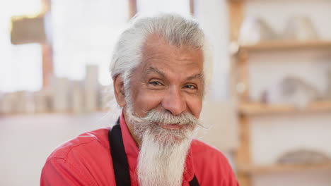 happy biracial potter with long beard looking on camera and smiling in pottery studio, slow motion