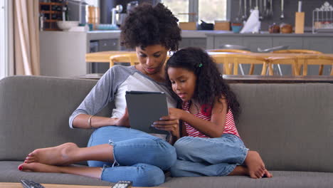 mother and daughter sit on sofa in lounge using digital tablet