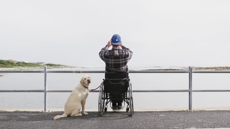 hombre en silla de ruedas tomando fotos del mar con su perro