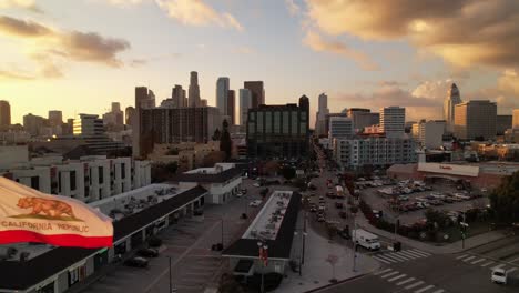 Los-Angeles-Skyline-Bei-Sonnenuntergang-Aus-Der-Luft-An-Der-Staatsflagge-Von-Kalifornien-Vorbei