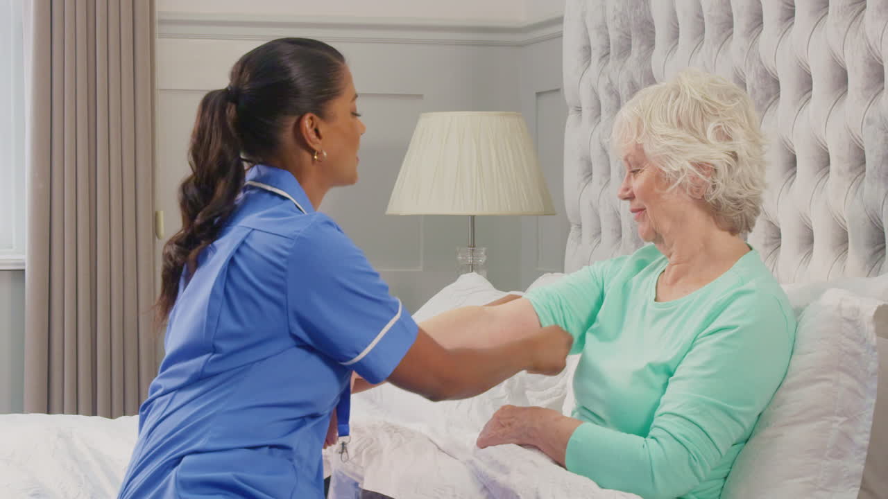 Senior Woman At Home In Bed Having Blood Pressure Taken By Female Care Worker In Uniform