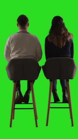 vertical video rear view studio shot of man and woman sitting on stools facing away from camera against green screen