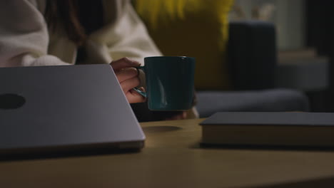 woman spending evening at home carrying hot drink and mobile phone sitting on sofa and opening laptop 1