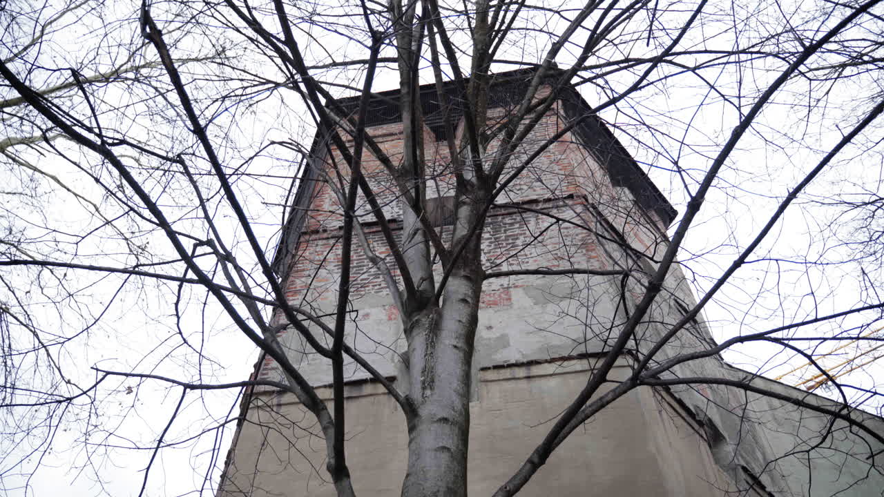 A historic medieval watchtower in Munich, Germany, partially obscured by leafless trees. The overcast sky and aged brick structure create a moody, winter atmosphere.