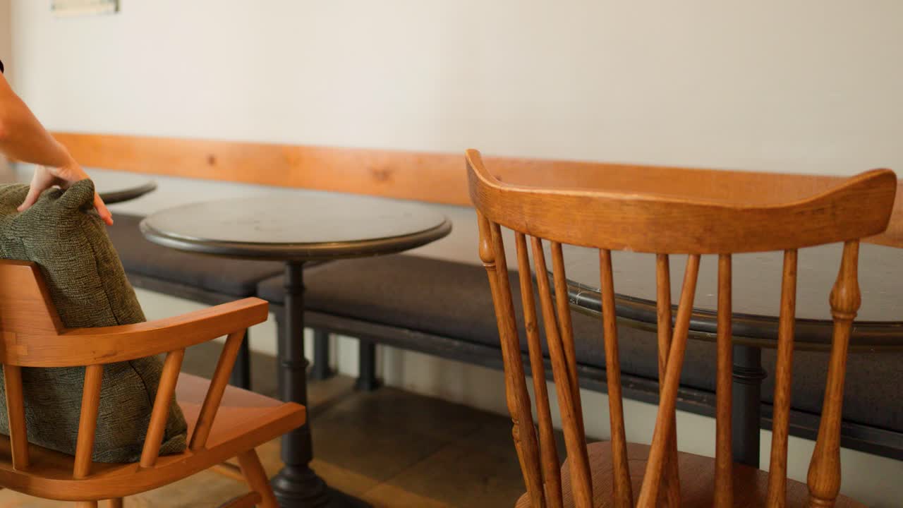 A person carefully places and adjusts green pillows on wooden chairs in a quiet cafe