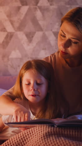 mommy teaches little girl to read following line with finger on bed near grey patterned wall in dark bedroom before going to sleep closeup slow motion