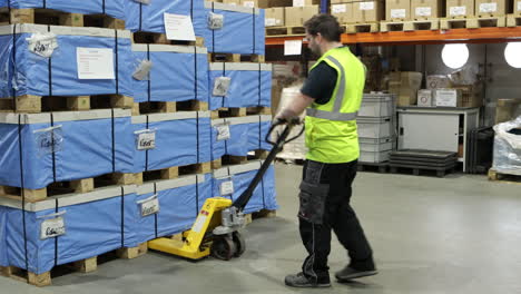 worker pulling pallet jack among stacked boxes in a busy warehouse, wearing safety vest