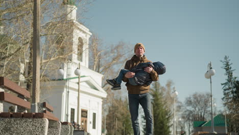 a front view of a father carrying his child while walking down a street, the father is dressed in a brown jacket and beanie, while the child is in a black puffy jacket, with white building behind