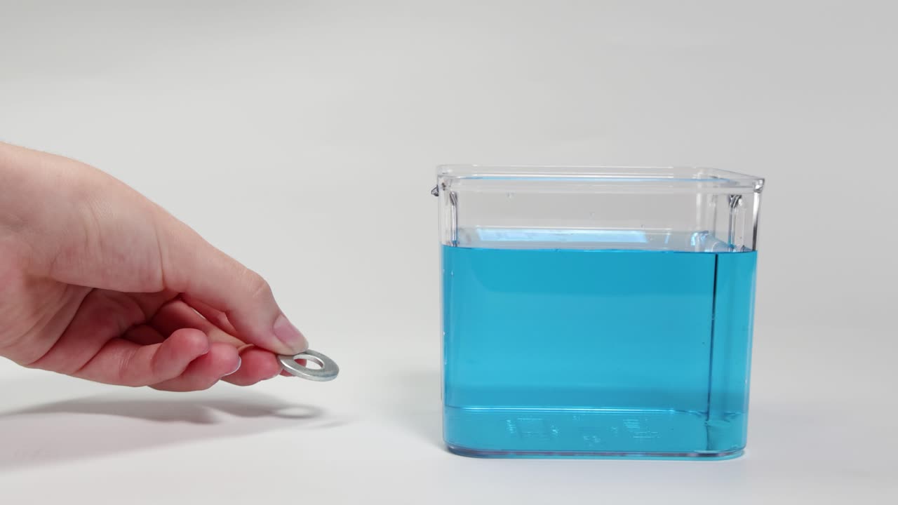 A hand drops a metal washer into a clear container with blue liquid, demonstrating buoyancy and sinking behavior