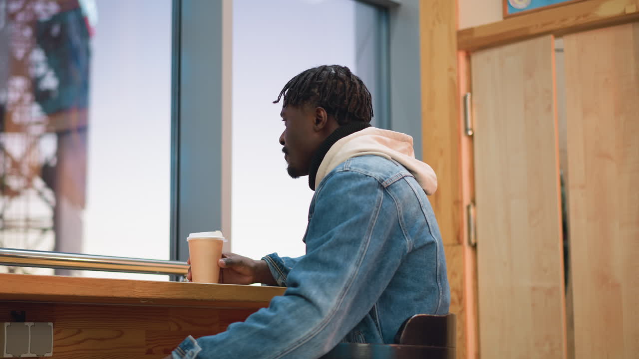 Young man in denim jacket and beige hoodie relaxes in cozy indoor cafe while sipping hot coffee from paper cup, gazing out large glass window as evening light fades behind city structure in soft sunset glow