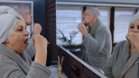 Senior-couple-grandmother-and-grandfather-brushing-teeth-and-looking-into-a-mirror-at-bathroom