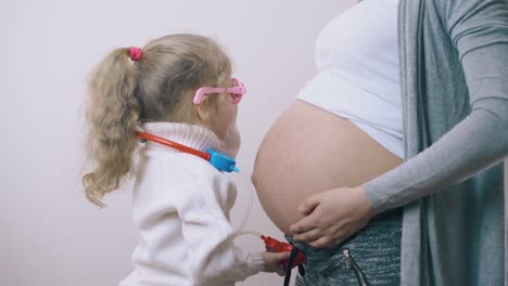 Kind-Mit-Spielzeugstethoskop-Hört-Schwangerer-Mama-Im-Zimmer-Zu