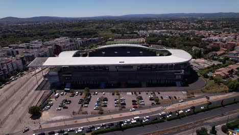 Voranschreitende-Luftaufnahme-Eines-Riesigen-Stadions-Im-Süden-Frankreichs-Mit-Einer-Stark-Befahrenen-Straße-Davor