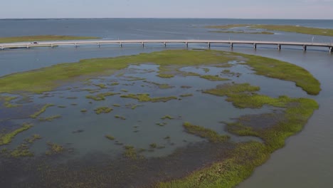 Lush-green-swamp-wetlands-below-concrete-bridge-crossing-water,-aerial-of-Chincoteague-Island-Virginia,-slow-motion