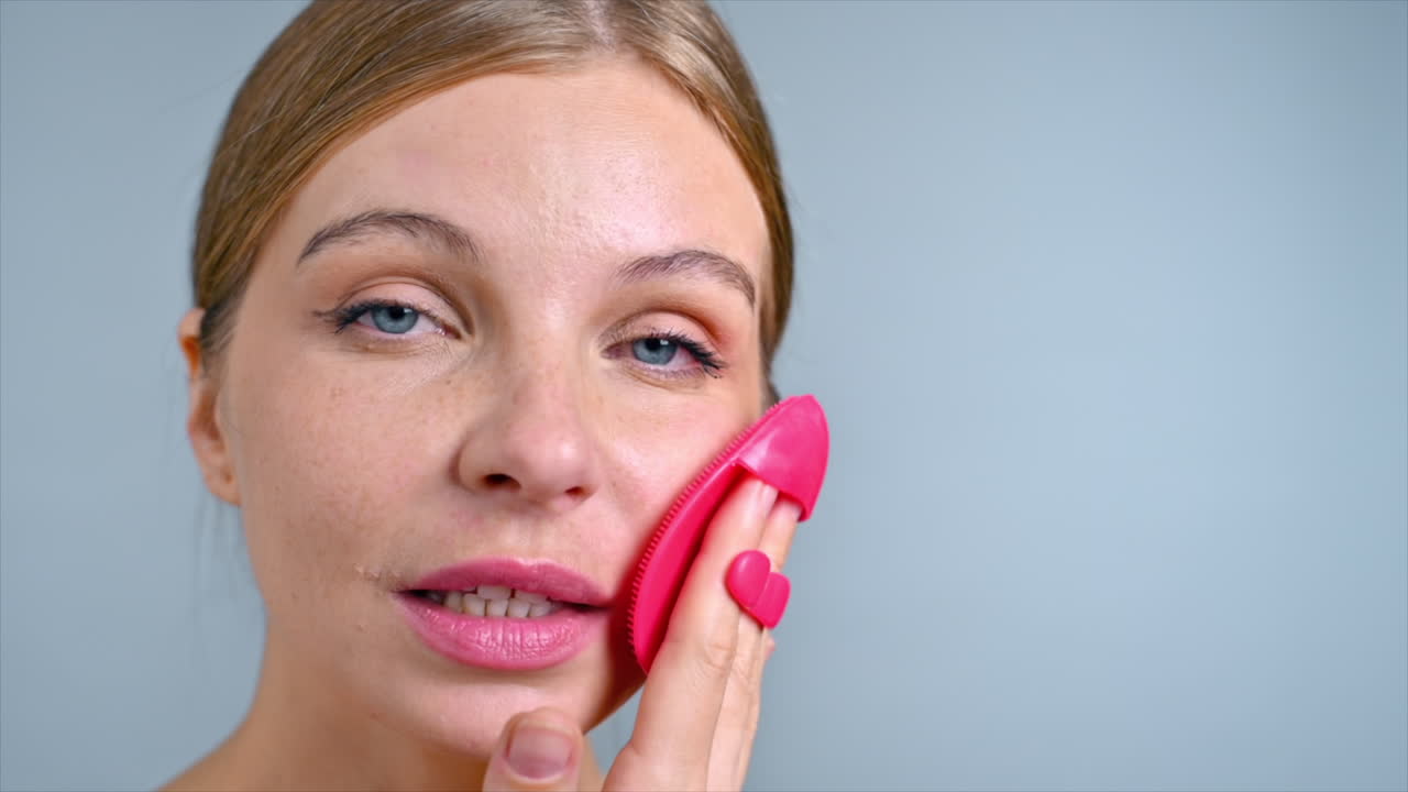 A young caucasian woman is doing a facial massage using face scrubber, looking into the camera. Slow motion