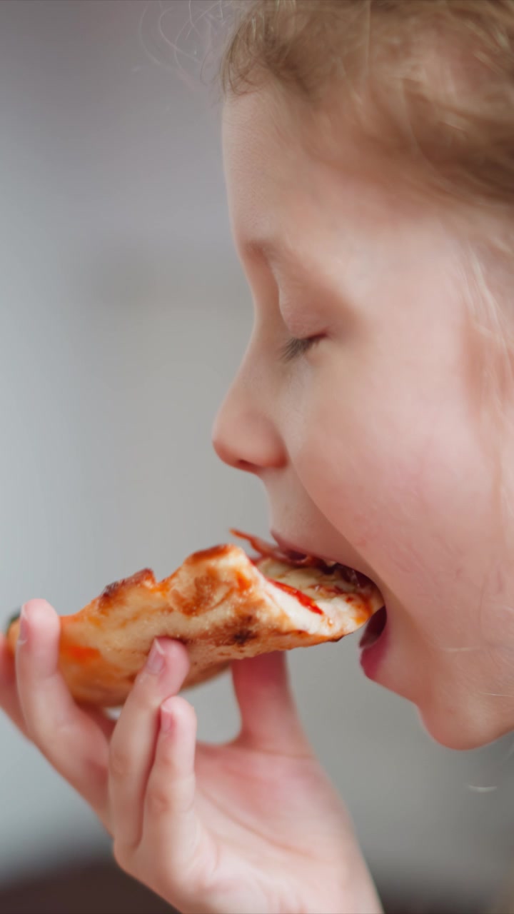 Close up of young girl eating pizza slice bringing it to mouth and taking bites. Hand holding food with soft lighting against plain background. Mouth movements show enjoyment during mealtime