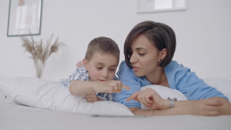 mamá e hijo mirando la pantalla de la tableta tendida en una cama blanca. juega juegos con tu hijo en tu tableta y mira videos divertidos.