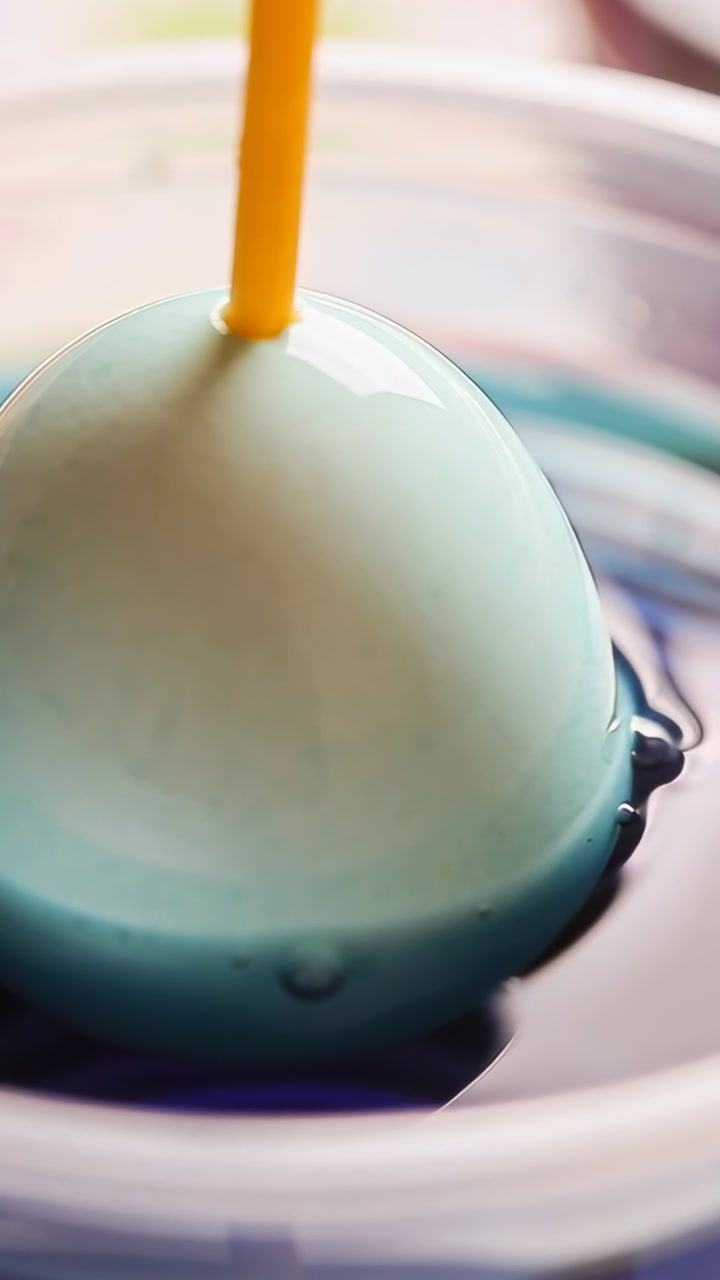 Yellow skewer lowering pale blue egg into craft bowl for dyeing, dye gradually coating glossy shell