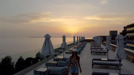 revealing shot of young woman strolling on poolside terrace at resort in sunset, greece