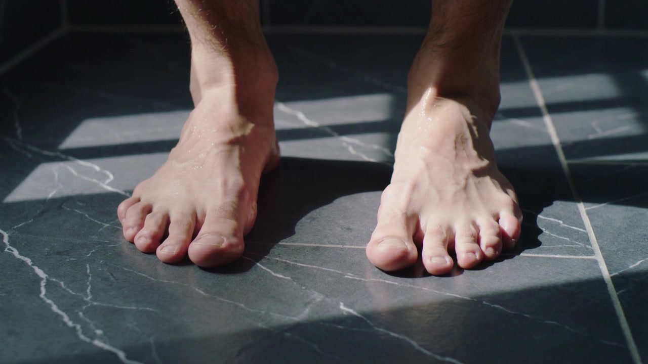 Piedi che si muovono asciugandosi su piastrelle scure di marmo del bagno mentre una fascia di luce solare cade, con una lucentezza bagnata.