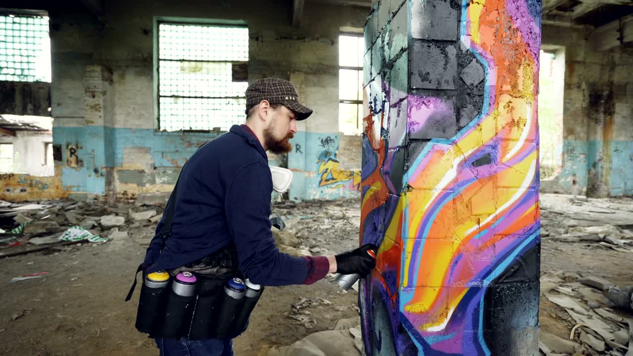 창의적인 수염이있는 남자가 손상된 벽과 더러운 바닥을 가진 오래된 건물 내부의 손상된 기둥에 그래피티를 칠하는 트업.