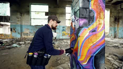 tilt-up shot of creative bearded guy painting graffiti on damaged column inside old building with damaged walls and dirty floor. creativity and young people concept.