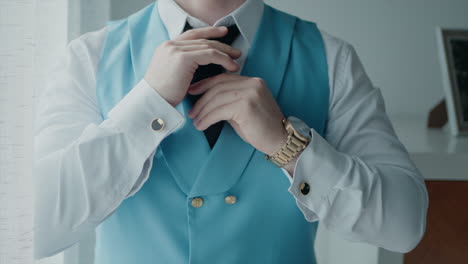 hombre ajustando su corbata, usando un chaleco azul claro sobre una camisa blanca, con un reloj en la muñeca, preparándose para un evento formal