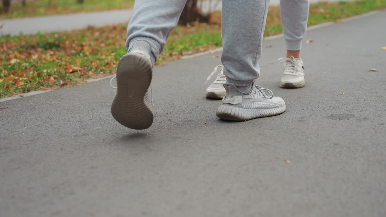 Lower angle view of two people walking up to each other on marked road surrounded by scattered autumn leaves and green grass in peaceful outdoor park setting