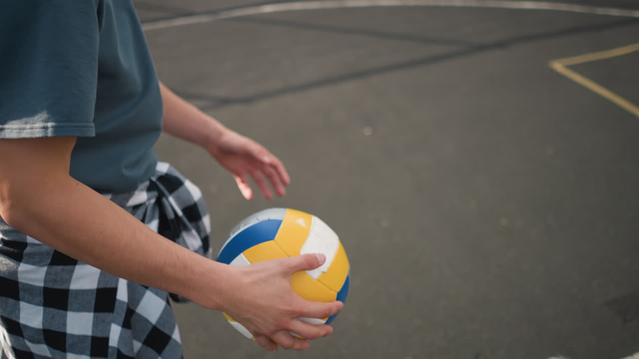 야외에서 공과 손에 초점을 맞추고 볼리볼을 뛰고 있는 허리를 둘러싼 카레드 셔츠를 입은 사람의 측면 뷰의 클로즈업