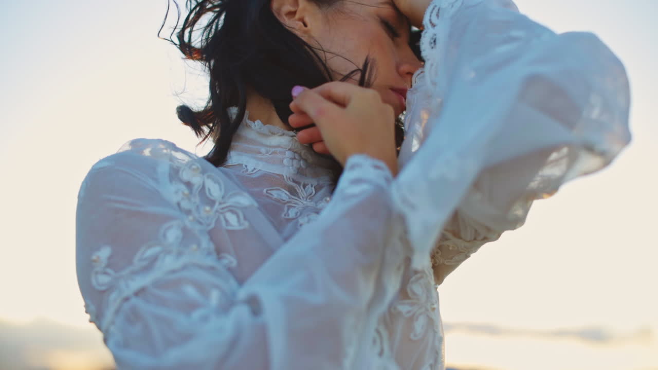 Long-haired woman in white dress enjoying wind in her hair. Lady stands outside in the rays of setting sun.
