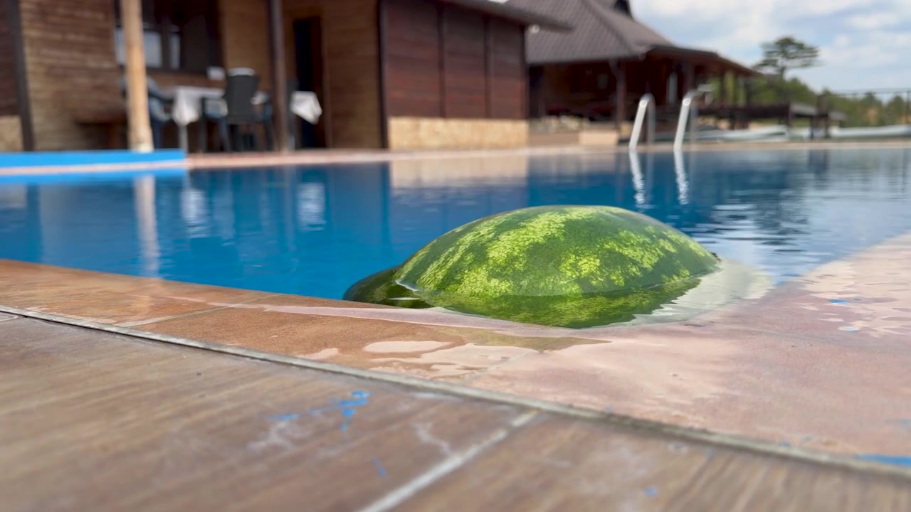 A striped watermelon bobs against the edge of a blue swimming pool in close up, with shallow focus revealing wet tan tiles, a metal ladder, and a wooden lodge beyond the water.