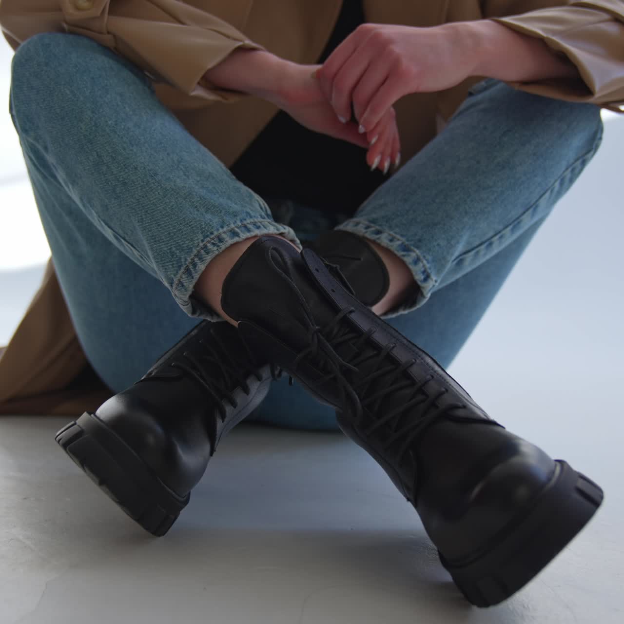 Girl in jeans and black army boots sits on the floor. Lady crosses her legs in front of camera demonstrating stylish footwear