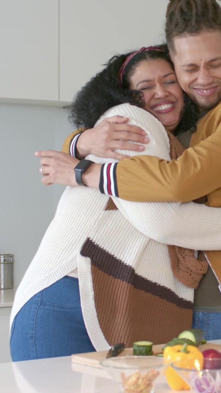 Video verticale: Uomo con cappotto color senape che taglia, si ferma mentre una donna gli mette una mano addosso e lo abbraccia vicino a una ciotola da cucina.