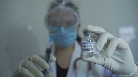 front-view-of-lady-doctor-wearing-apron-and-stethoscope-holding-covid-19-vaccine-in-one-hand-and-syringe-in-another