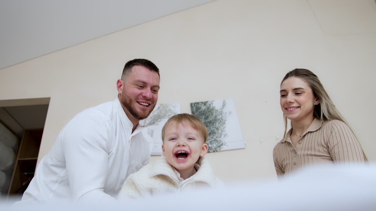 Father sitting on the sofa waves and tosses his little cheerful baby boy. Loving parents kiss their adorable son on the cheek. Low angle view.