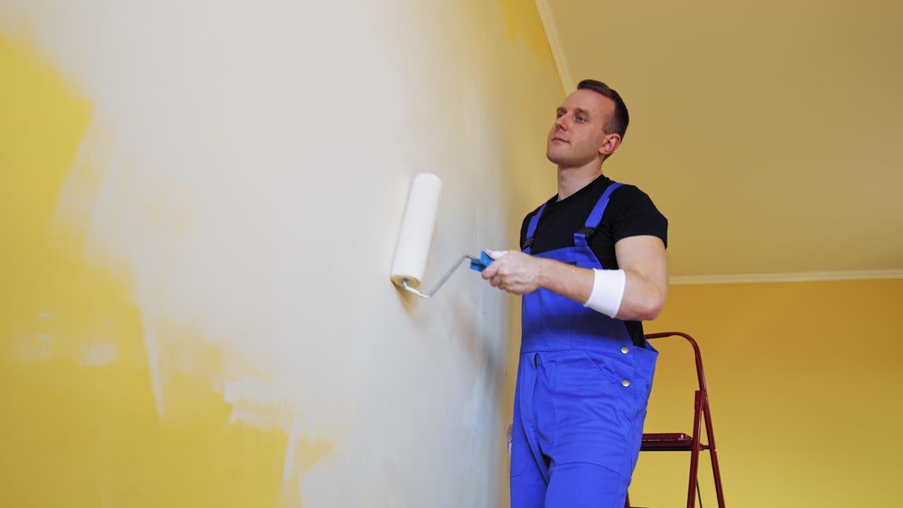 Painter man painting wall with a roller. Portrait of a young worker in overalls doing makeover with new color in the room. Professional painting services.
