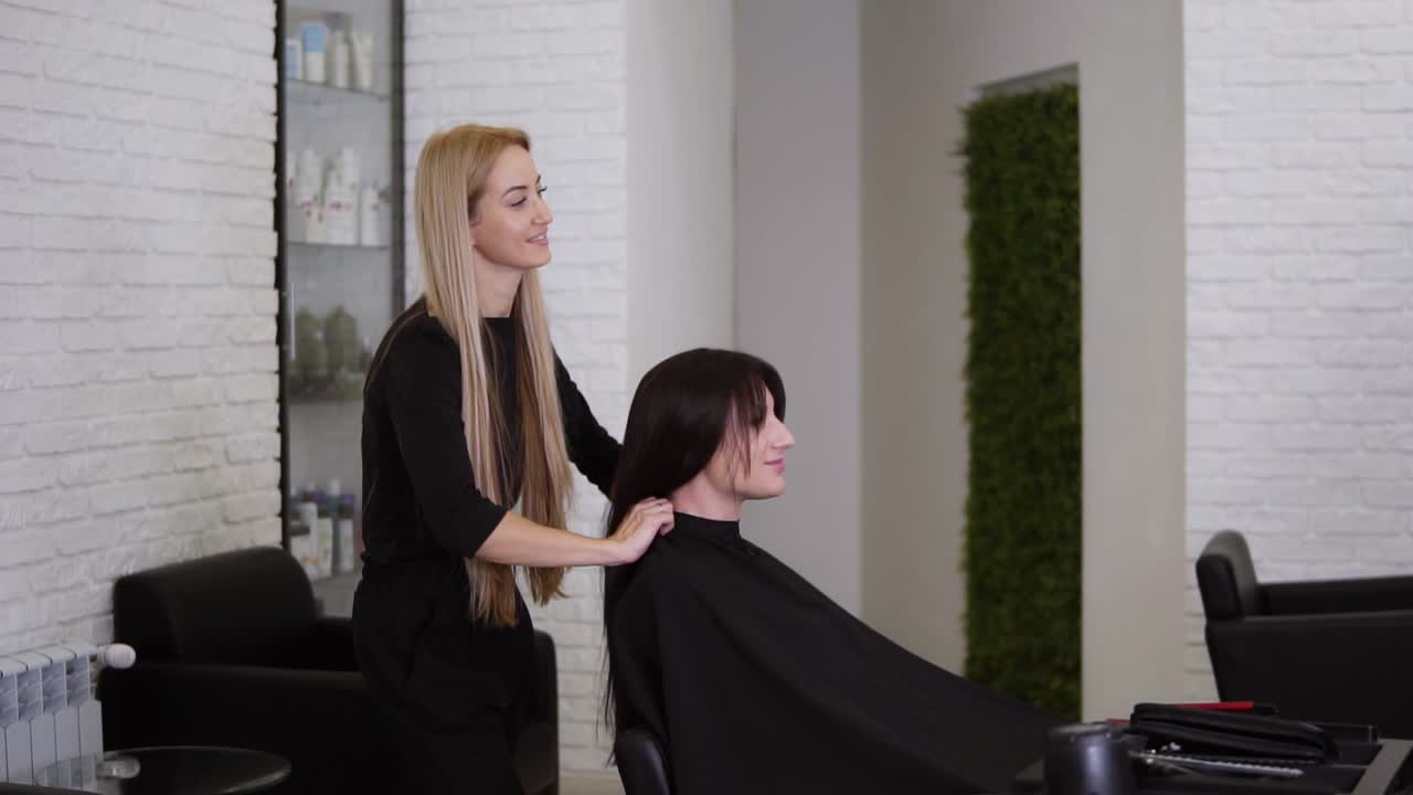 Female hairdresser has just finished working on her female client with long, straight brown hair. Woman is smiling and glad about her new hair style. Hairdresser removing protective cape. Side view
