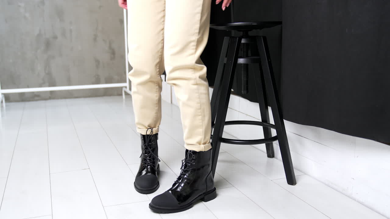 Unknown woman comes up to a high stool and sits down. Model demonstrates stylish boots from combined leather. White backdrop.