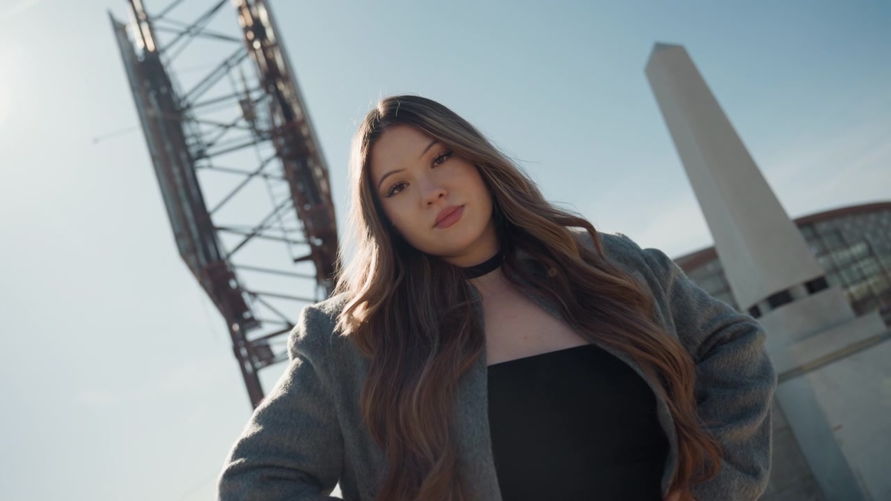 Ritratto di donna asiatica in controluce dal basso, bella direttrice creativa che guarda verso l'alto con lunghi capelli che catturano i riflessi, sagoma di torre d'acciaio e obelisco sullo sfondo, toni caldi cinematografici e che trasmette forza