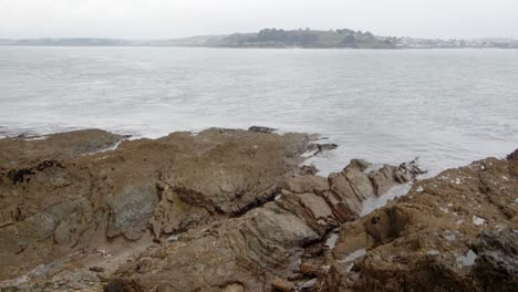 Wide-Shot-of-waves,-lapping-on-jagged,-seaweed-rocks-with-St-Mawes-in-the-background