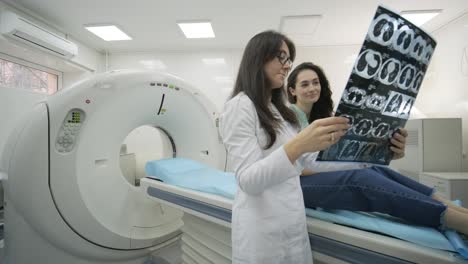 woman doctor radiologist explains good results of ct scanning for young female patient, showing the snapshot with images, observing and analyzing ct scan in modern clinic beside modern ct scanner