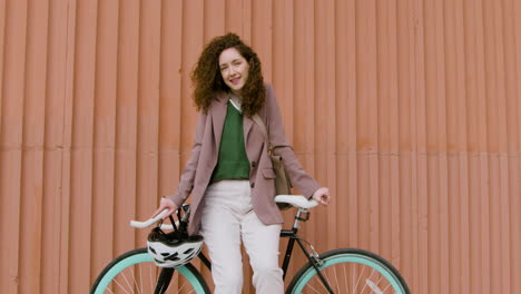 smiling curly girl in formal clothes looking at the camera while leaning on bike in front of a prefab metal building 1