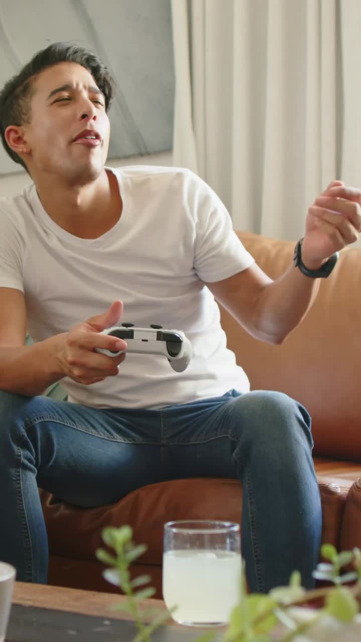 Vertical video: Reacting to game man playing controller on sofa at home wearing white tee and jeans