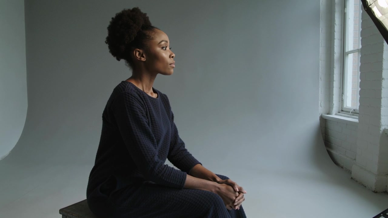 Raising chin model in dark dress, turning toward window light adjusting pose on bench in studio